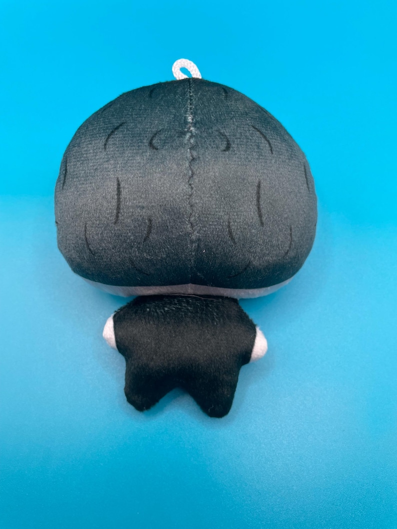 May include: A plush doll with a black head and body, and white accents on the feet. The doll has a white loop at the top for hanging. The background is a solid light blue.