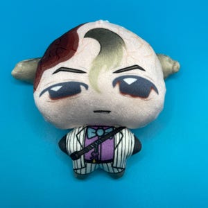 May include: A plush doll with a light skin tone, featuring a stylized face with dark eyes and a distinctive hairstyle. The doll is dressed in a purple shirt, a white pinstripe suit, and a black strap across the chest. The background is a solid blue.