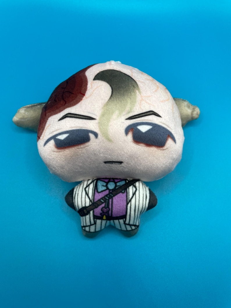 May include: A plush doll with a light skin tone, featuring a stylized face with dark eyes and a distinctive hairstyle. The doll is dressed in a purple shirt, a white pinstripe suit, and a black strap across the chest. The background is a solid blue.