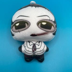 May include: A plush doll with a white head and body, featuring a stylized face with large, dark eyes and a small red mouth. The doll's outfit includes a white shirt with a black tie and black pants. The doll is set against a solid blue background.