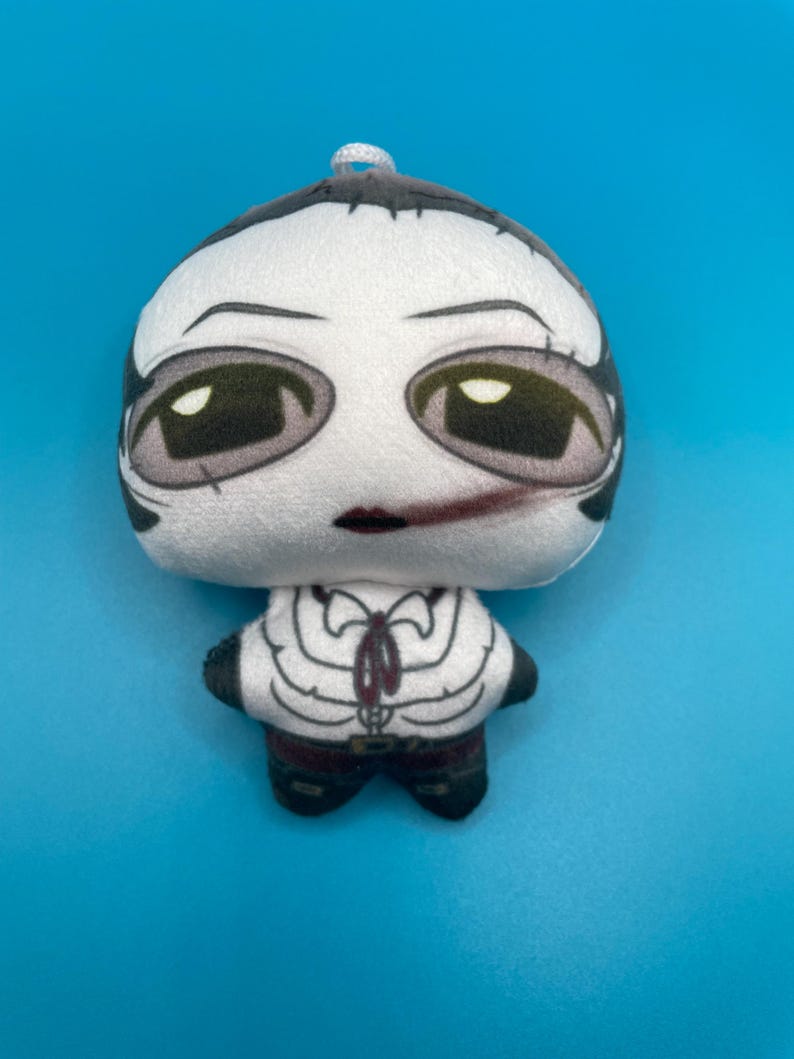 May include: A plush doll with a white head and body, featuring a stylized face with large, dark eyes and a small red mouth. The doll's outfit includes a white shirt with a black tie and black pants. The doll is set against a solid blue background.