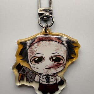 May include: A clear acrylic keychain featuring a cartoon character with a stitched head, bloodied face, and dark hair. The character holds a small bird with a top hat. The keychain has a silver clasp.