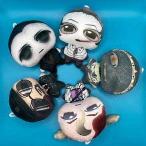 May include: A collection of plush dolls with various character designs. The dolls feature detailed facial expressions and are made of soft fabric. The color palette includes black, white, and shades of beige and brown, set against a bright blue background.