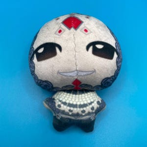 May include: A plush doll with a round head and a small body. The doll's head is off-white with black eyes and a small smile. The doll has a red diamond shape on its forehead and a patterned collar.