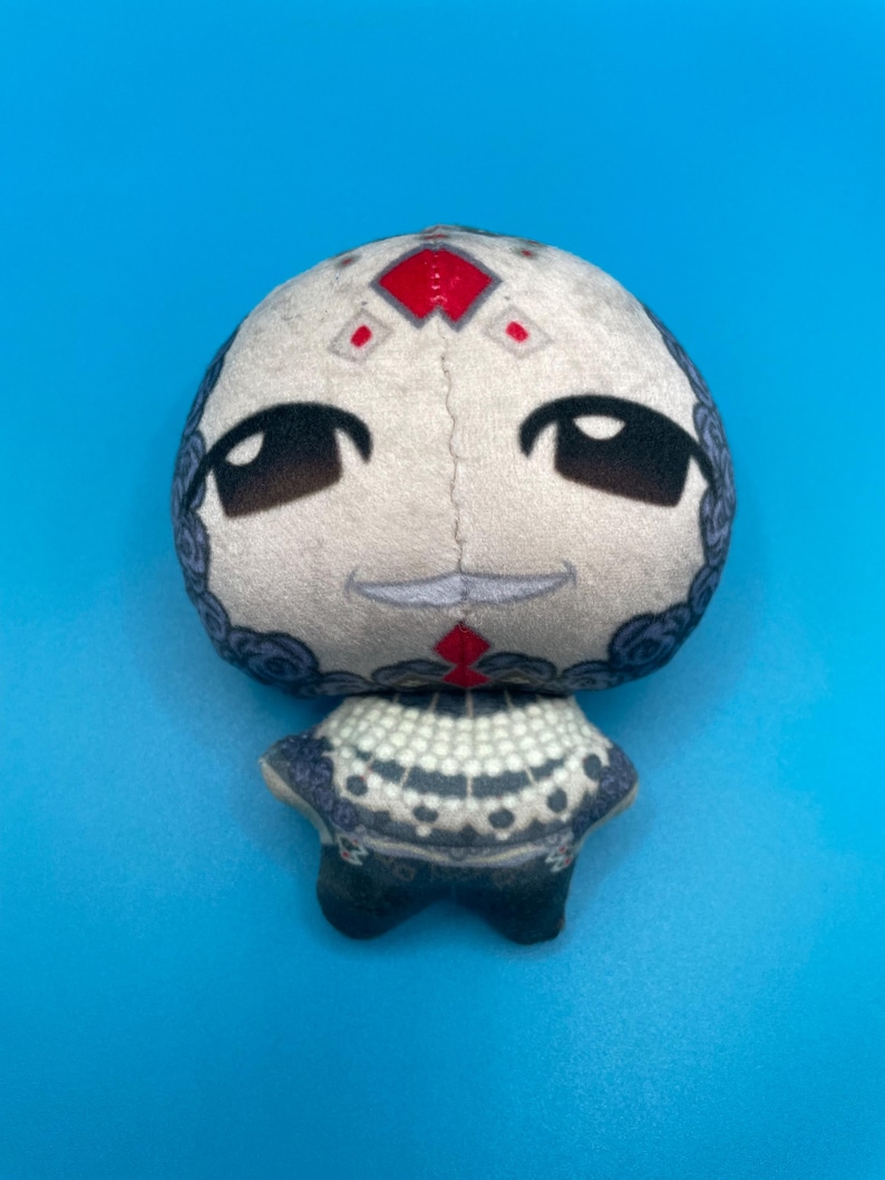 May include: A plush doll with a round head and a small body. The doll's head is off-white with black eyes and a small smile. The doll has a red diamond shape on its forehead and a patterned collar.