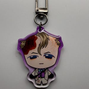 May include: A purple-edged keychain featuring a cartoon character with a stylized design. The character has light skin, blue eyes, and a formal outfit. The character's hair is light brown, and there is a red and brown design on the head.