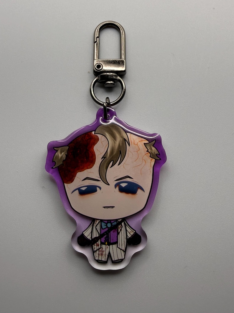May include: A purple-edged keychain featuring a cartoon character with a stylized design. The character has light skin, blue eyes, and a formal outfit. The character's hair is light brown, and there is a red and brown design on the head.