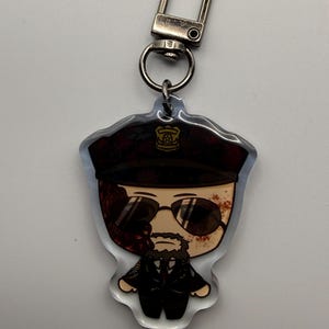 May include: A clear acrylic keychain featuring a cartoon illustration of a man in a police uniform with sunglasses. The keychain has a silver-colored clasp and ring for attaching to keys or bags.