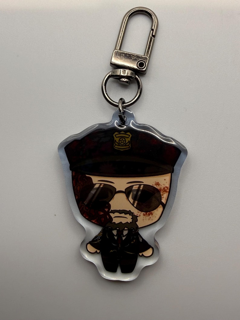 May include: A clear acrylic keychain featuring a cartoon illustration of a man in a police uniform with sunglasses. The keychain has a silver-colored clasp and ring for attaching to keys or bags.