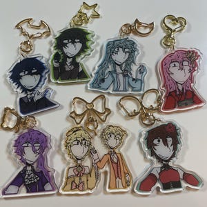 Può includere: Otto portachiavi di personaggi colorati con anelli in metallo dorato. I personaggi sono disegnati in stile cartone animato e presentano diversi colori di capelli, vestiti ed espressioni.