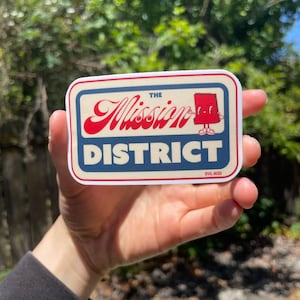 Op de afbeelding: Een rechthoekige sticker met een crèmekleurige achtergrond en een rood en blauw ontwerp. De tekst "The Mission District" wordt weergegeven in een vintage lettertype. Een klein stripfiguur staat aan de rechterkant van de sticker. De sticker wordt in een hand gehouden.