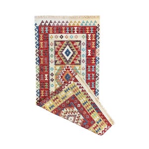 Puede incluir: Una alfombra rectangular plegada con un diseño geométrico. La alfombra presenta un diseño central de diamante en blanco, azul y amarillo, rodeado de bordes rojos y crema. Tiene un borde con flecos y una mezcla de colores.