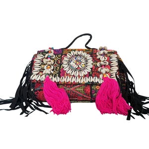 May include: A rectangular handbag featuring a multi-colored embroidered fabric base with black fringe along the sides. The front flap is decorated with white cowrie shells arranged in circular and linear patterns, accented by two large bright pink tassels.