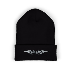 May include: Black beanie hat featuring a white tribal design on the folded brim. The hat is made of a soft material, suitable for colder weather. The tribal design is symmetrical, adding a unique touch to the hat.