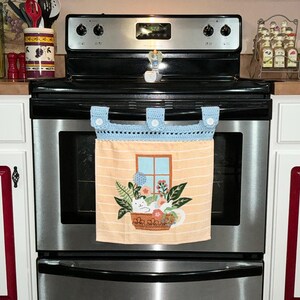 May include: A light orange kitchen towel with a blue crocheted top, hanging on a stainless steel oven. The towel features a window design with a cat, flowers, and greenery. The towel is held in place by three white buttons.