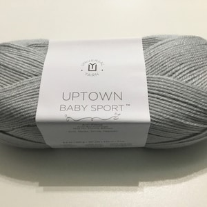 May include: A skein of light gray Uptown Baby Sport yarn by Universal Yarn. The label reads "Uptown Baby Sport" and includes the Universal Yarn logo. The yarn is anti-pilling and suitable for knitting and crochet projects. The skein weighs 3.5 oz (100 g).
