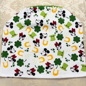 May include: White fabric kitchen tea cozy with a green, yellow, and red pattern of Mickey Mouse and Minnie Mouse heads, shamrocks, horseshoes, and leprechaun hats.