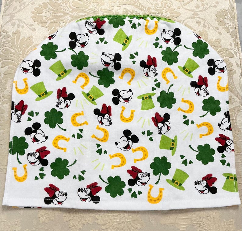 May include: White fabric kitchen tea cozy with a green, yellow, and red pattern of Mickey Mouse and Minnie Mouse heads, shamrocks, horseshoes, and leprechaun hats.