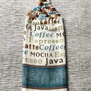May include: A white kitchen towel with a crocheted brown and blue hanging loop. The towel has a blue and brown border with a design of coffee cups and coffee pots. The center of the towel has a white background with the words "so Lafie C", "HA Java Esp", "Coffee MO", "Espresso", "atte Coffeer", "e MOCHA ESH", and "resso Java" printed in brown, blue, and green.