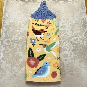 May include: A yellow kitchen towel with a blue crocheted hanging loop. The towel features a colorful floral pattern with three birds, including a hummingbird, a bluebird, and a brown bird.