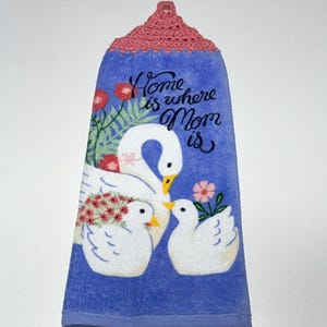 May include: Blue kitchen towel with a crocheted pink top. The towel features a white swan with two baby swans and the text "Home is where Mom is".