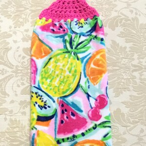 May include: A pink crocheted hanging loop attached to a white kitchen towel with a colorful fruit print. The print features pineapples, lemons, oranges, watermelon, cherries, and kiwi.