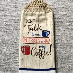 May include: A beige kitchen towel with a crocheted hanging loop. The towel has a woodgrain background with the text "Don't Talk to me... Unless I've Had My Coffee!" and two illustrations of coffee mugs.