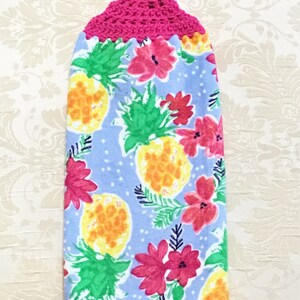 May include: A hanging kitchen towel with a bright pink crocheted top. The towel features a light blue background with a pattern of yellow pineapples, pink flowers, and green leaves. The towel is designed for kitchen use.