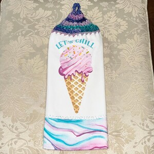 May include: White hanging kitchen towel with a crocheted top in shades of blue, purple, and green. The towel features a large pink ice cream cone graphic with the words "LET'S CHILL" in blue. The bottom has a watercolor design.