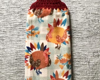 May include: A red crocheted kitchen towel holder with a button closure. The towel is white with a blue and white gingham pattern and features a colorful turkey print.