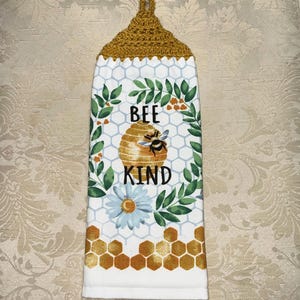 May include: A white kitchen towel with a crochet top, featuring a bee-themed design. The towel has a honeycomb pattern, a bee, and the words "BEE KIND". Green leaves and a blue flower surround the bee and the text.