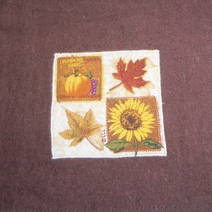 May include: A square fabric patch with a white background and a brown border. The patch is divided into four sections, each featuring a different autumnal image. The top left section shows a pumpkin with the text "Pumpkin Farm". The top right section shows a red maple leaf. The bottom left section shows a brown leaf. The bottom right section shows a sunflower.