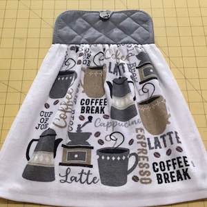 May include: White hanging kitchen towel with a gray quilted top and a button closure. The towel features coffee-themed graphics, including coffee cups, coffee pots, and coffee beans, with the words "Coffee Break" and "Latte".