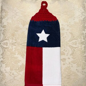 May include: A hanging kitchen towel featuring the Texas state flag design. The top portion is red crochet, transitioning to a navy blue section with a white star. The bottom half is divided into red and white vertical stripes.