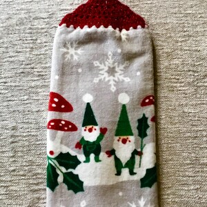 May include: A gray kitchen towel with a red crocheted hanging loop. The towel features a winter scene with two gnomes, holly, mushrooms, and snowflakes.