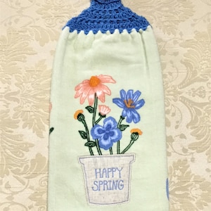 May include: A light green kitchen towel with a crocheted blue hanging loop and a button. The towel features a floral design with blue, pink, and yellow flowers in a pot with the text "Happy Spring".