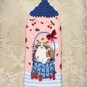 May include: A pink kitchen towel with a blue crochet top and loop. The towel features a cat in a blue basket with strawberries and cherries. The bottom has blue and white stripes.
