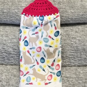 May include: A white hanging kitchen towel featuring a repeating pattern of beige cat silhouettes, yellow flowers, pink and blue tulips, and colorful Easter eggs. The top of the towel has a bright pink crocheted topper with a small loop for hanging.