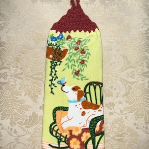 May include: A yellow kitchen towel with a burgundy crochet top. The towel features a dog sitting in a chair, with a butterfly and hanging plants. The dog has brown spots and a blue collar.