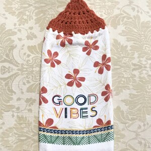 May include: A white hanging kitchen towel with a crocheted rust-colored top. The towel features red hibiscus flowers and the words "GOOD VIBES" in colorful letters. A decorative border with geometric patterns accents the bottom.