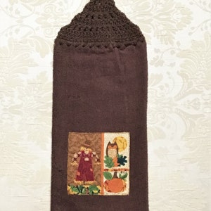 May include: Brown kitchen towel with a crocheted hanging loop. The towel features a patchwork design with a scarecrow, owl, and pumpkin.