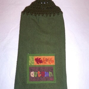 May include: Green kitchen towel with a crocheted loop at the top. The towel has a rectangular patch with an embroidered design of autumn leaves and the word "autumn" in colorful letters.