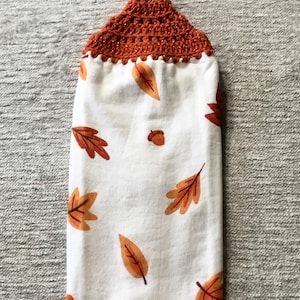 May include: A white kitchen towel with a brown crocheted hanging loop and a pattern of orange and brown leaves.