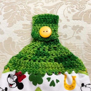 May include: Green crocheted kitchen towel with a yellow button. The towel has a white background with a green shamrock and horseshoe design. The towel is decorated with a cartoon image of Mickey Mouse.