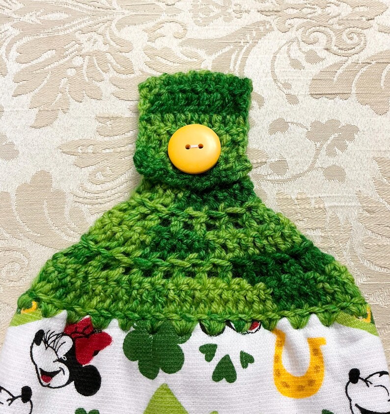 May include: Green crocheted kitchen towel with a yellow button. The towel has a white background with a green shamrock and horseshoe design. The towel is decorated with a cartoon image of Mickey Mouse.