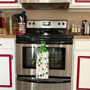May include: A green, white, and red kitchen towel with a Mickey Mouse and shamrock design hangs on a silver oven door handle.