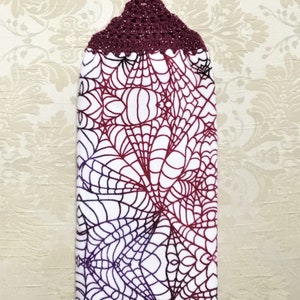May include: A white kitchen towel with a purple and red spiderweb design. The towel has a burgundy crocheted hanging loop.