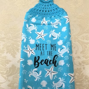 May include: A blue kitchen towel with a crocheted hanging loop and a white button. The towel has a white and black print of seashells, starfish, and crabs. The text "Meet me at the beach" is printed in black on the towel.