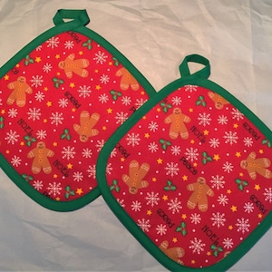 May include: Two red and green Christmas pot holders with a gingerbread man and snowflake pattern. The pot holders have the words "Peace" and "Noel" on them.