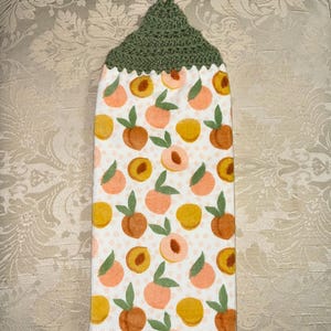 May include: A hanging kitchen towel with a green crocheted top and a white body. The body of the towel is patterned with peaches in shades of orange, yellow, and pink, with green leaves. The peaches are scattered across a white background.